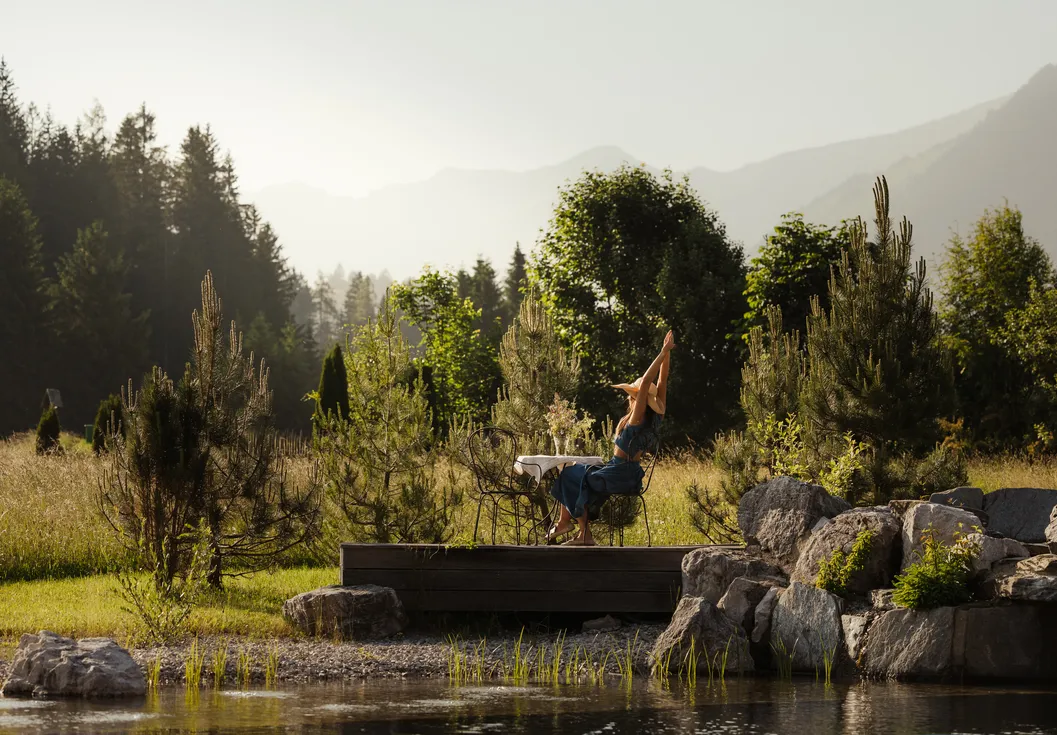 Woman raising her arms joyfully on a wooden terrace by the nature pond with alpine panorama