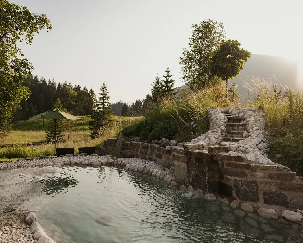 Natural stone pool with cascading waterfall, reed grass and a green parasol in evening light