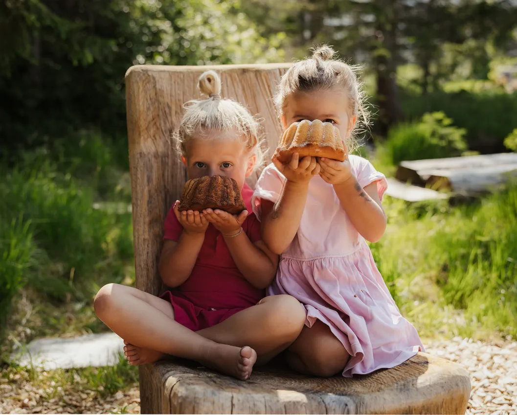 Two blonde girls sitting barefoot on a wooden chair holding up a Bundt cake