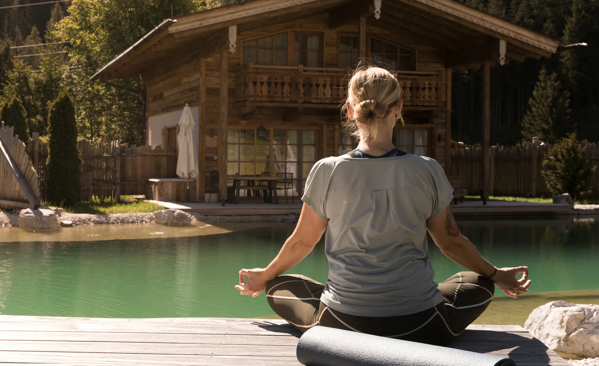 Frau meditiert im Lotus-Sitz auf Holzdeck am türkisfarbenen Naturpool bei Chalet