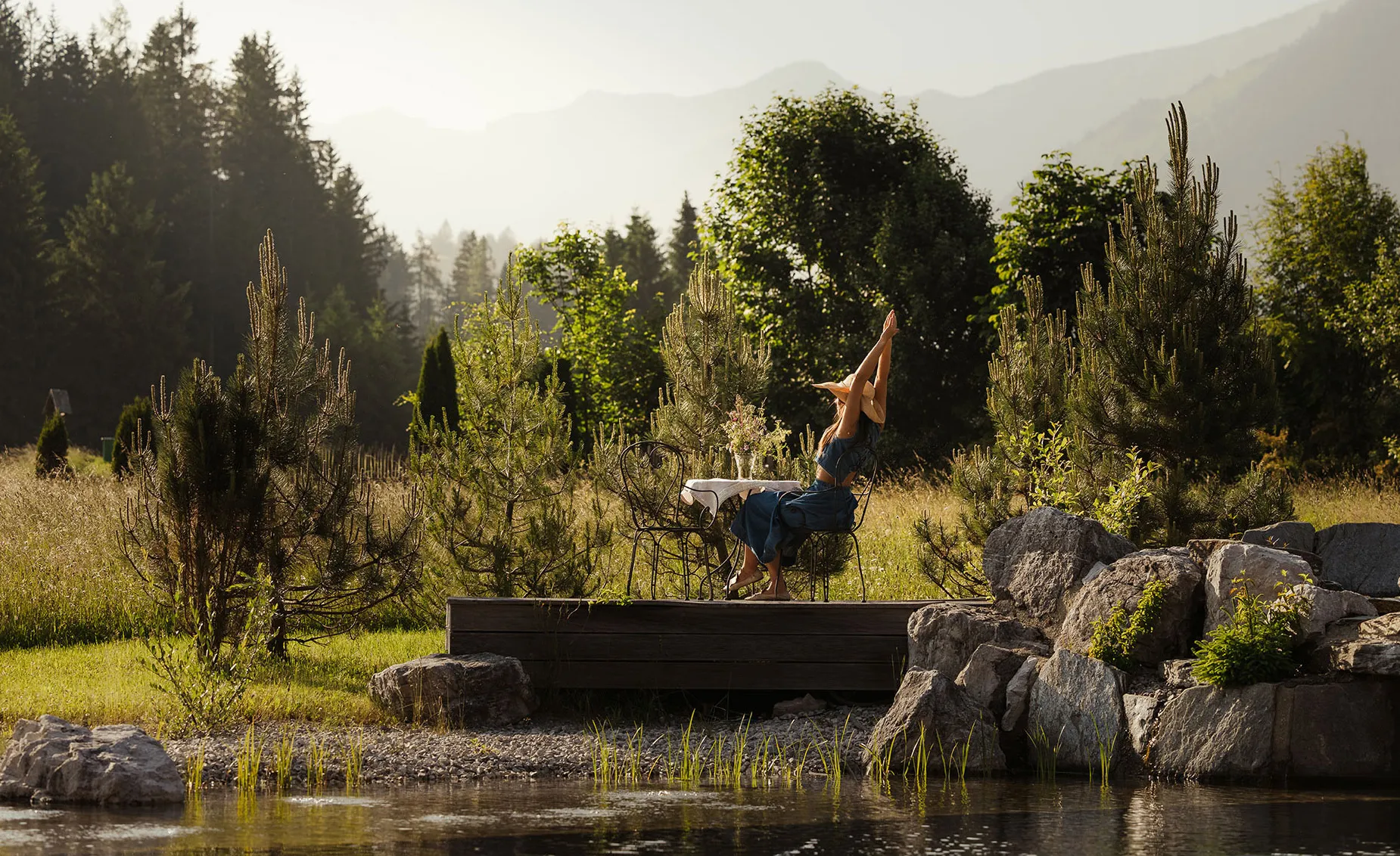 Frau macht Yoga-Kopfstandpose auf Holzdeck am Naturteich mit Alpenpanorama hinter sich