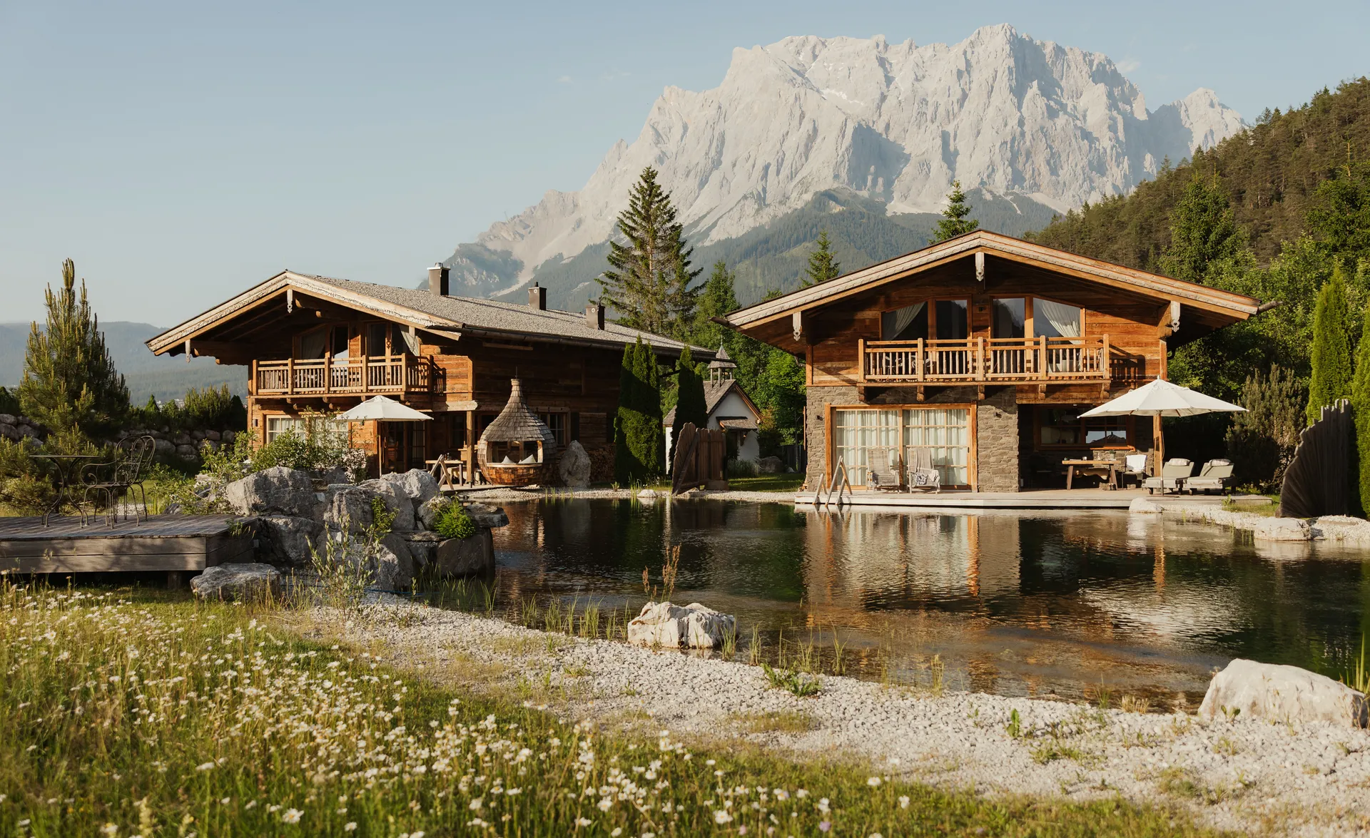 Zwei Holzchalets mit Balkonen spiegeln sich im Naturteich vor der Zugspitze