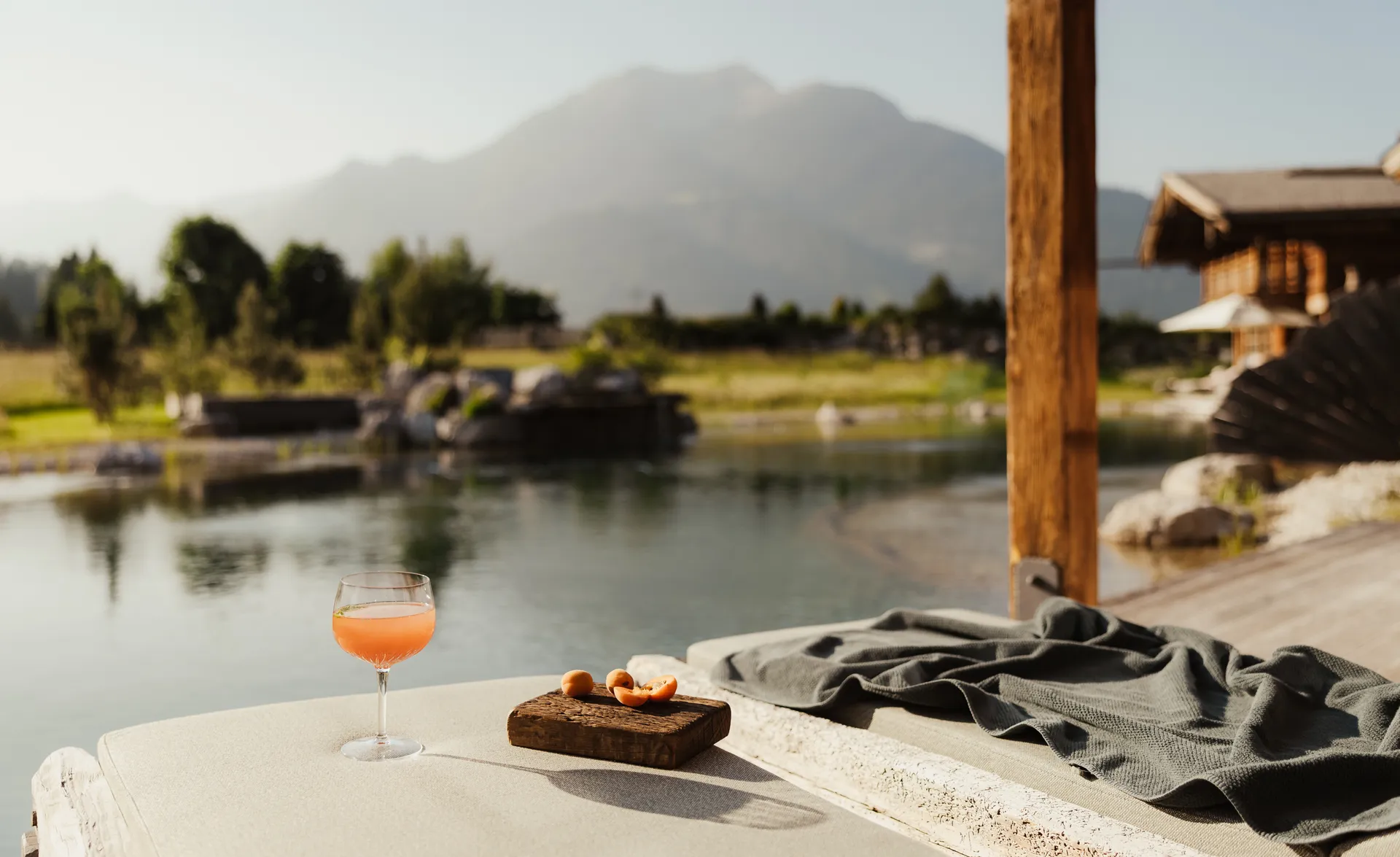 Aperitif-Glas mit Orangencocktail an der Liegefläche am Kristallsee mit Bergpanorama