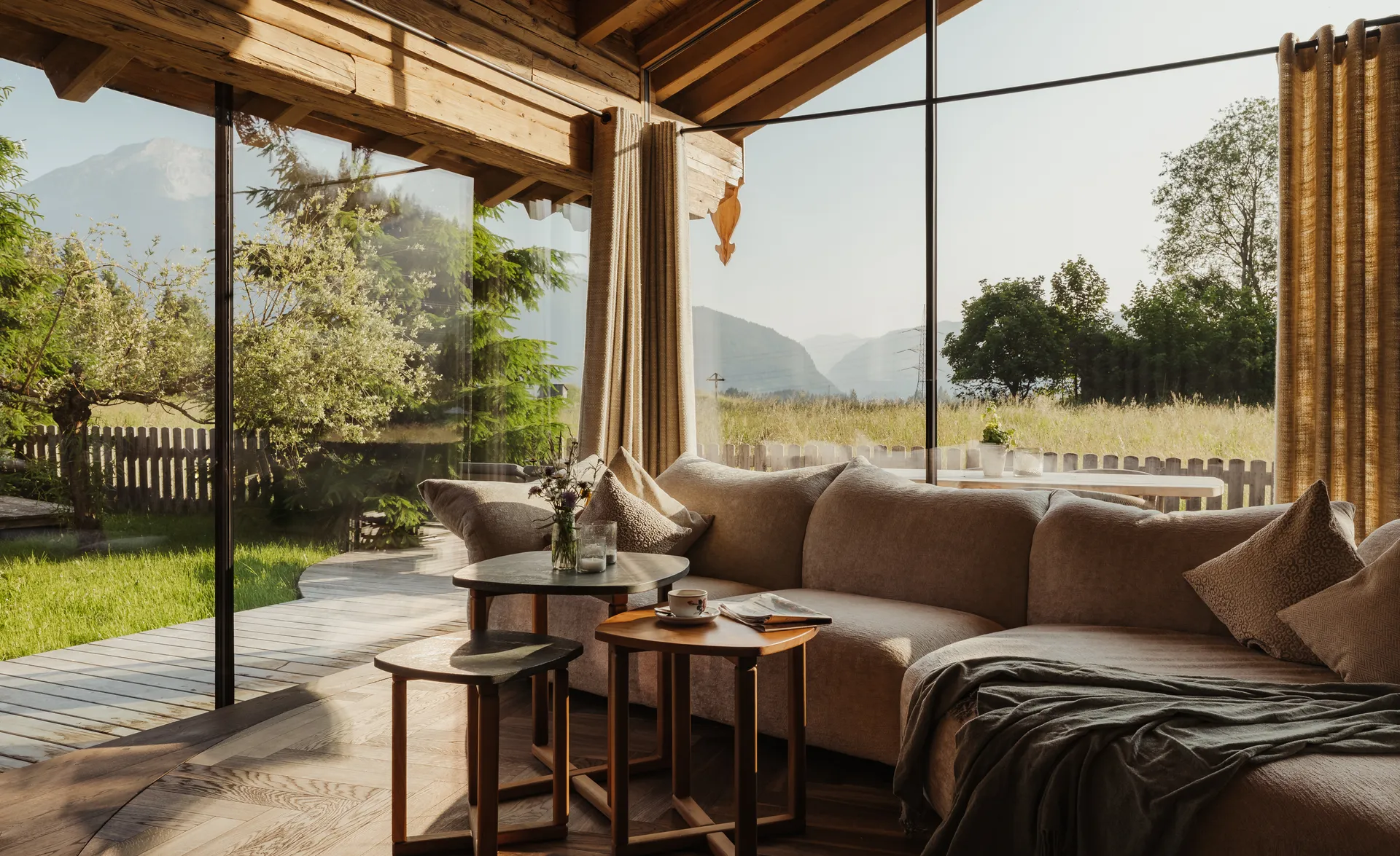 Chalet-Wohnraum mit Glasfront, Ecksofa, Holzbalken und Blick auf Wiese und Gipfel