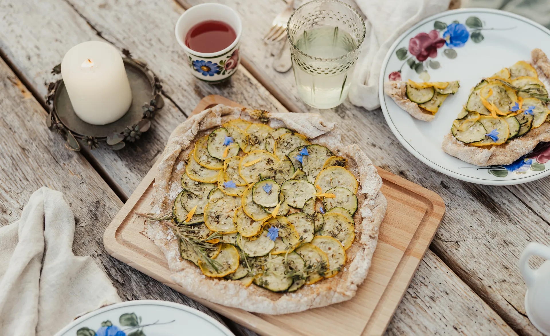 Aufsicht auf hausgemachte Zucchini-Galette auf Holzbrett mit Kerze und Getränken