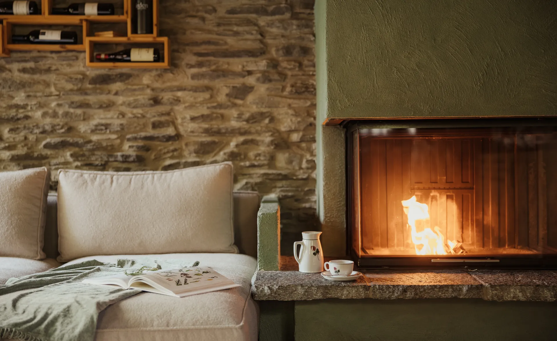Gemütlicher Kaminofen in Naturstein mit beigem Sofa und Weinregal im Chalet