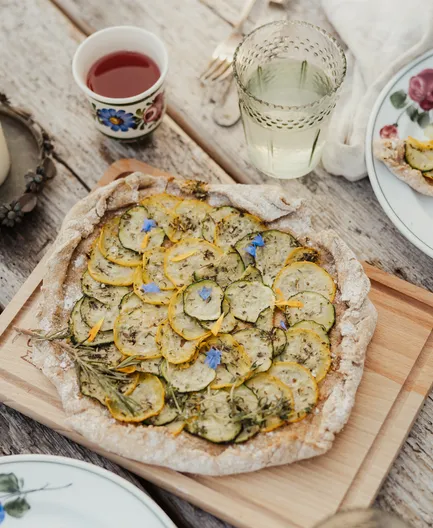 Aufsicht auf hausgemachte Zucchini-Galette auf Holzbrett mit Kerze und Getränken