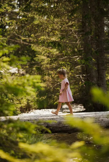 Kleines Mädchen in rosa Kleid balanciert barfuß auf Baumstamm über Gebirgsbach