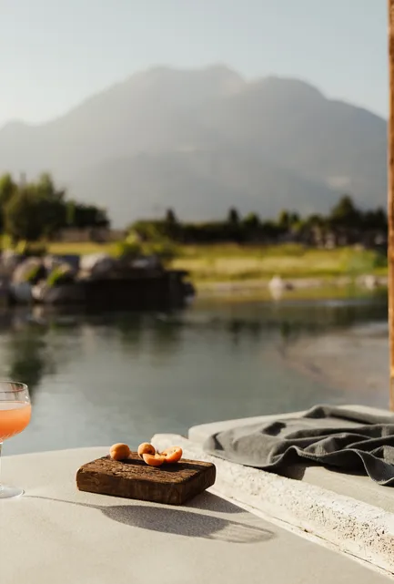 Aperitif-Glas mit Orangencocktail an der Liegefläche am Kristallsee mit Bergpanorama