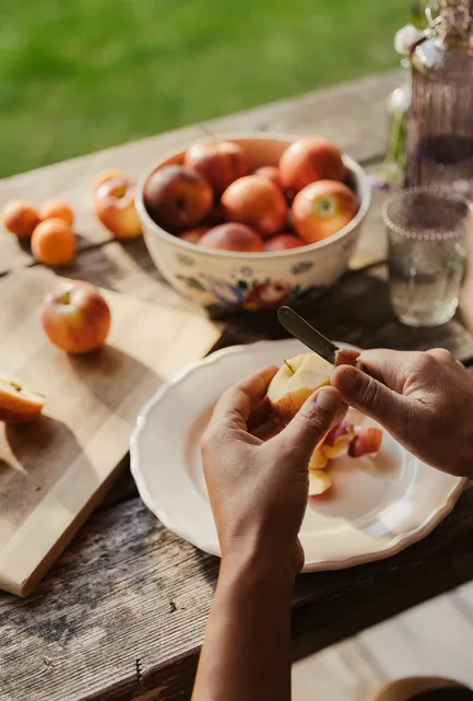 Hands peeling an apple with a knife over a plate at a rustic outdoor table