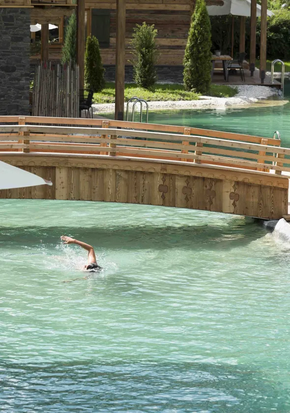 Person schwimmt Kraulstil im türkisfarbenen Naturpool neben Holzsteg des Chalets