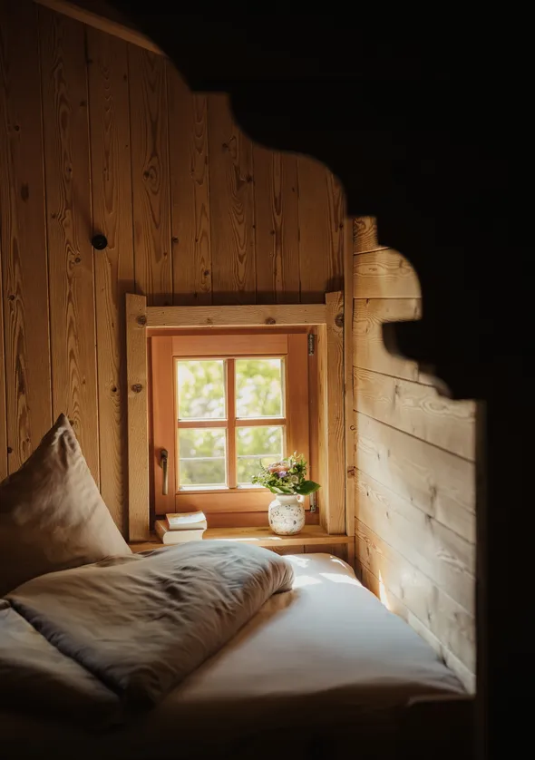 Gemütliche Chalet-Schlafnische mit kleinem Holzfenster, Wildblumen und weichem Leinenstoff