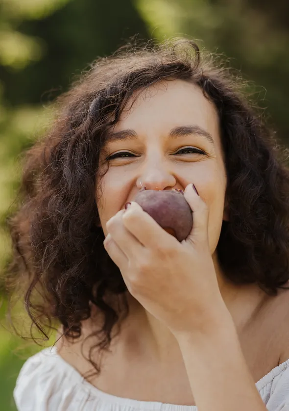 Curly-haired woman biting into a ripe plum and laughing outdoors in the summer