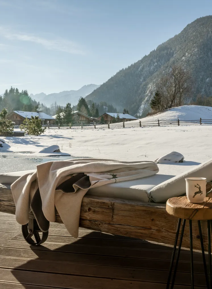 Holzliege mit Polster und Decke auf sonniger Chalet-Terrasse mit Alpenwinterpanorama