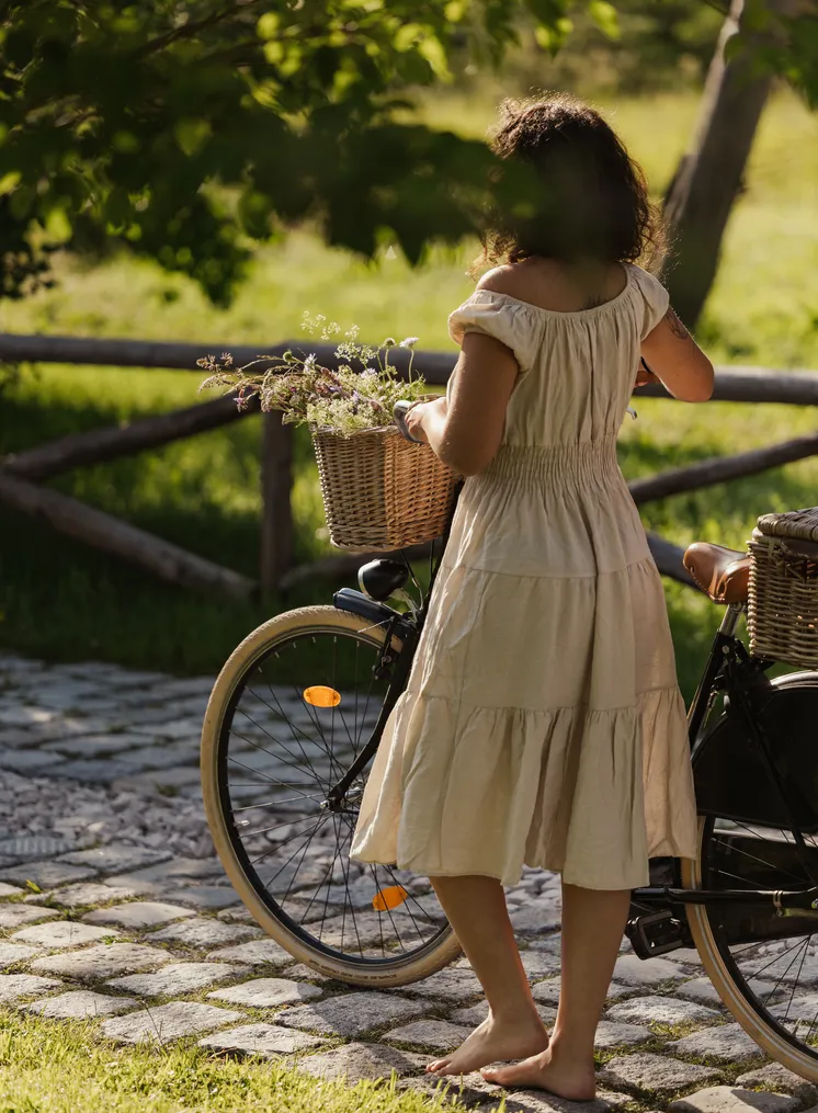 Frau im weißen Kleid steht barfuß mit Fahrrad und Wildblumenkorb auf Kopfsteinpflaster