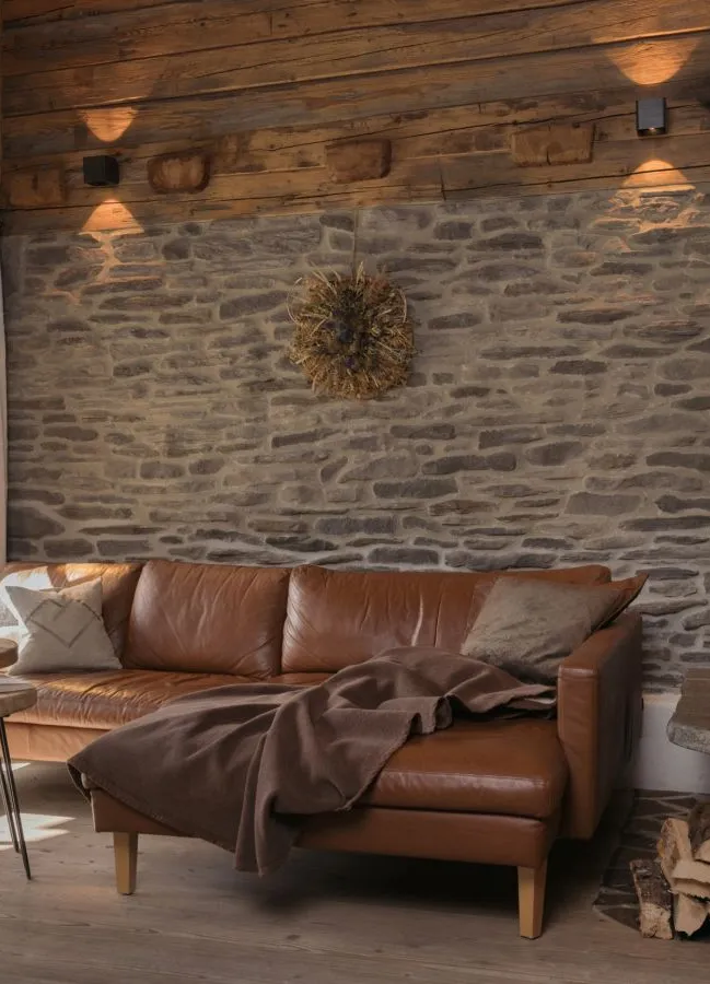 Chalet-Wohnzimmer mit braunem Ledersofa, Naturstein-Kaminofen und Holzbalkendecke