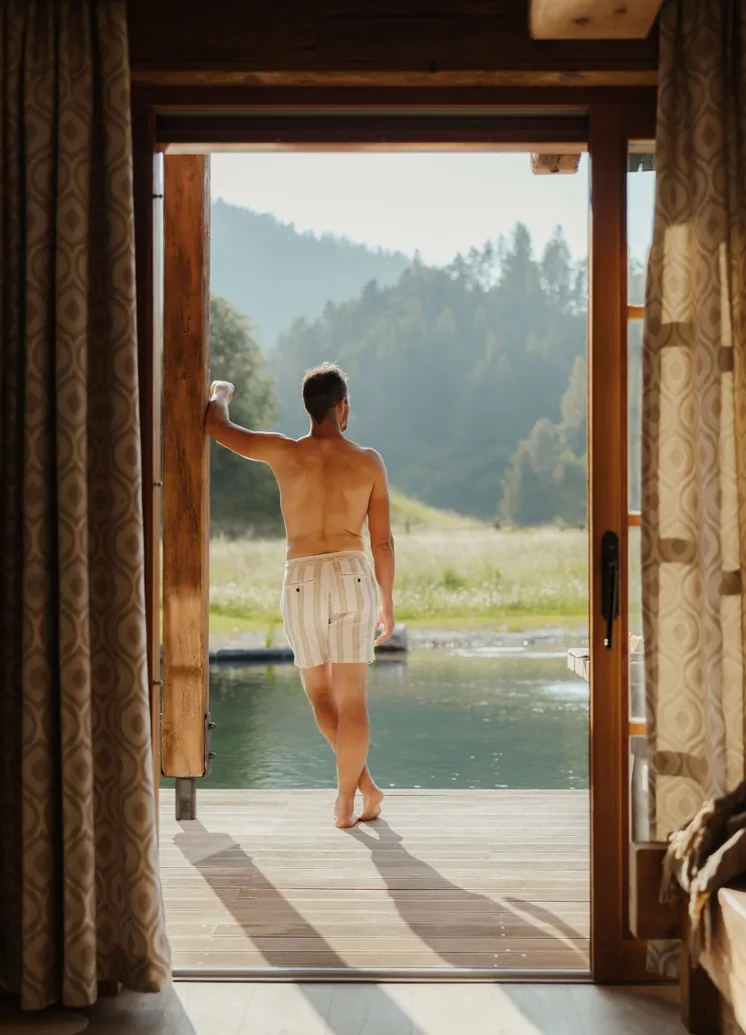 Mann steht in offenem Chalet-Rahmen und blickt auf Naturteich und Tiroler Bergwelt