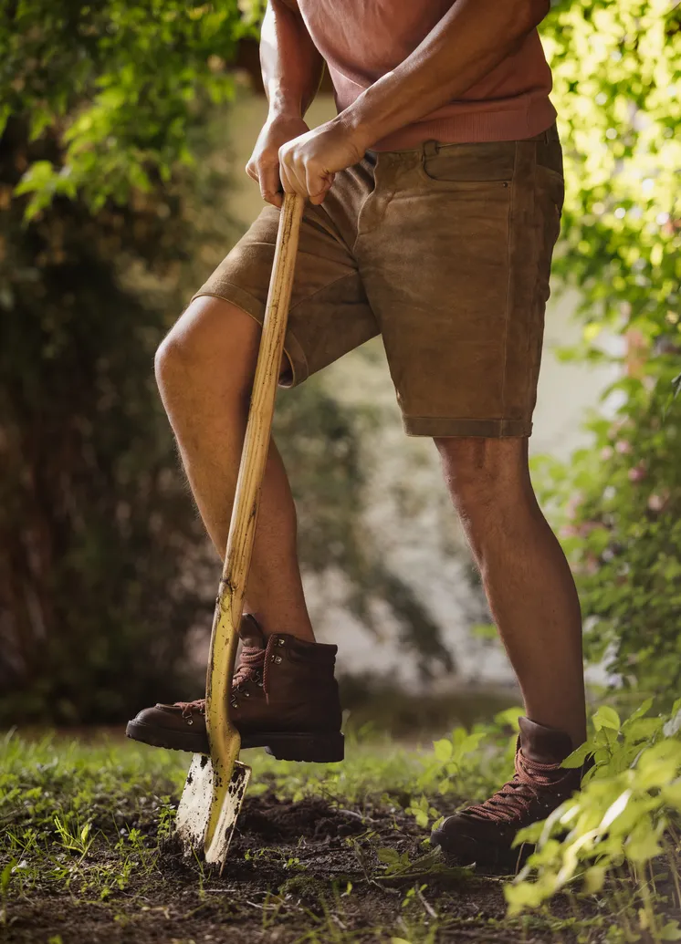 Mann in Lederhosen gräbt mit Holzspaten im Gartenbereich des LaPosch Resorts