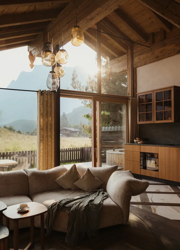Airy chalet living room with wooden roof truss, designer pendants and mountain view through the glass facade