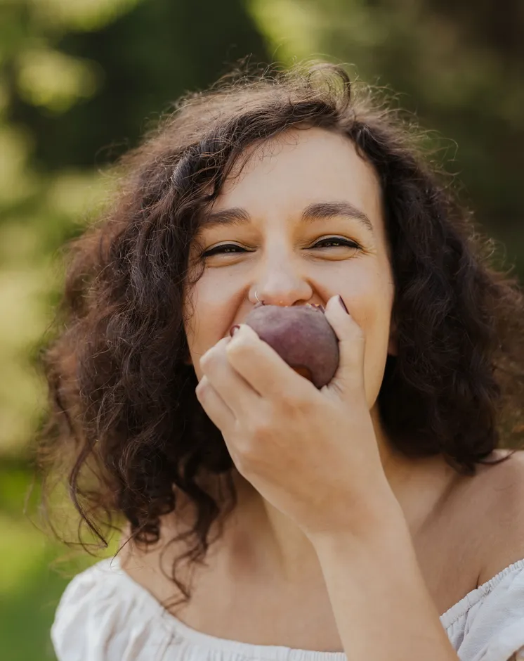 Smiling curly-haired woman laughing while biting into a ripe plum outdoors